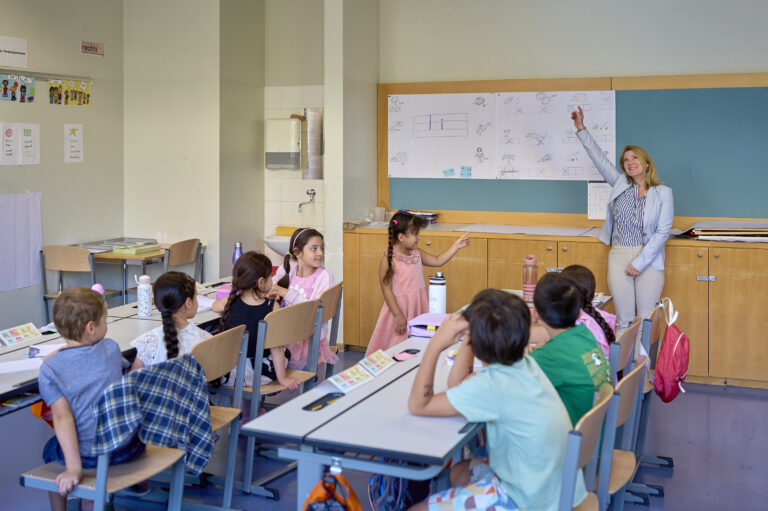 Kinder sitzen im Klassenzimmer, Vizebürgermeisterin zeigt auf